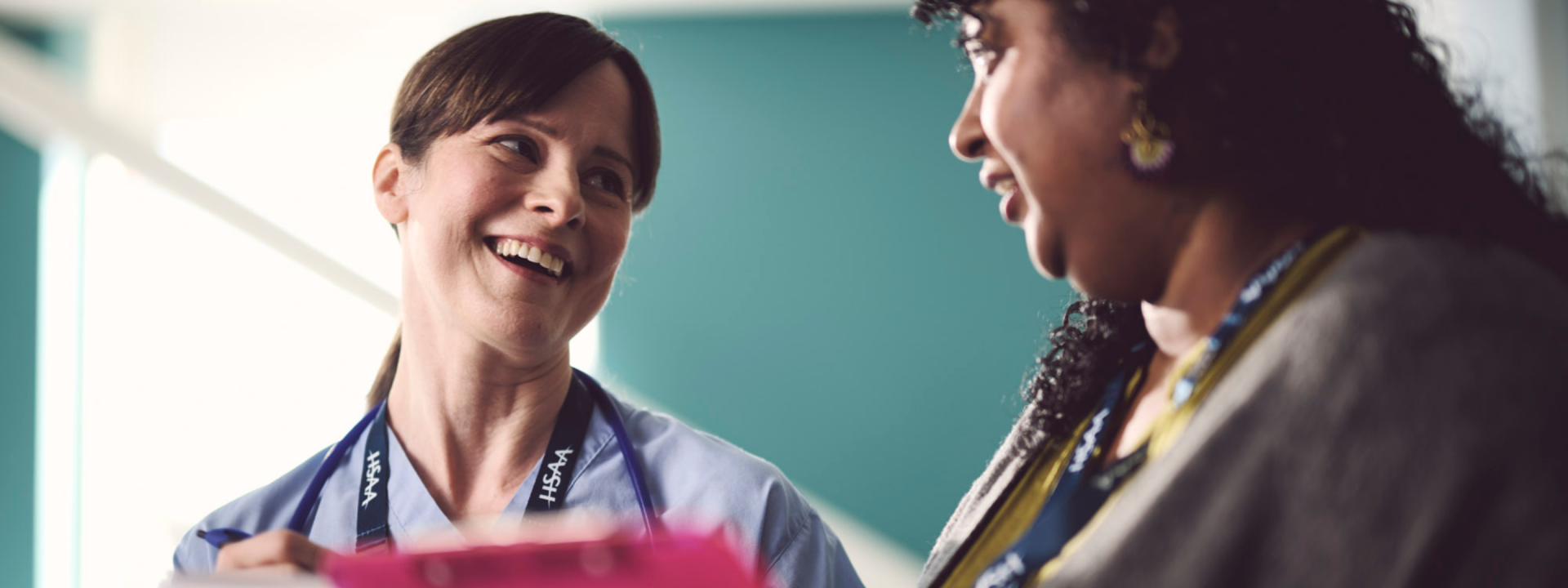 A white woman in scrubs and a brown woman in casual clothes talking to each other and smiling. Both are holding union paperwork.