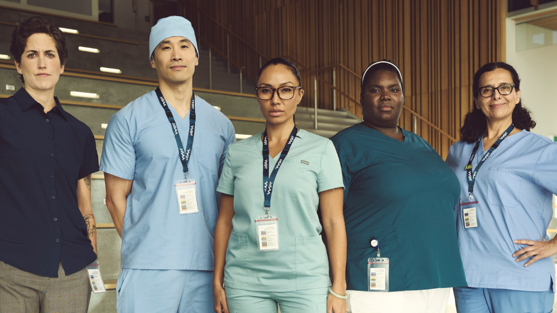 Five health care professionals in work appropriate attire stand in an open space, looking seriously at the camera. 