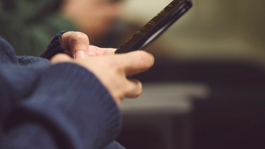 Two hands holding a mobile phone, with another pair of hands holding a phone out of focus in the background of a waiting room.