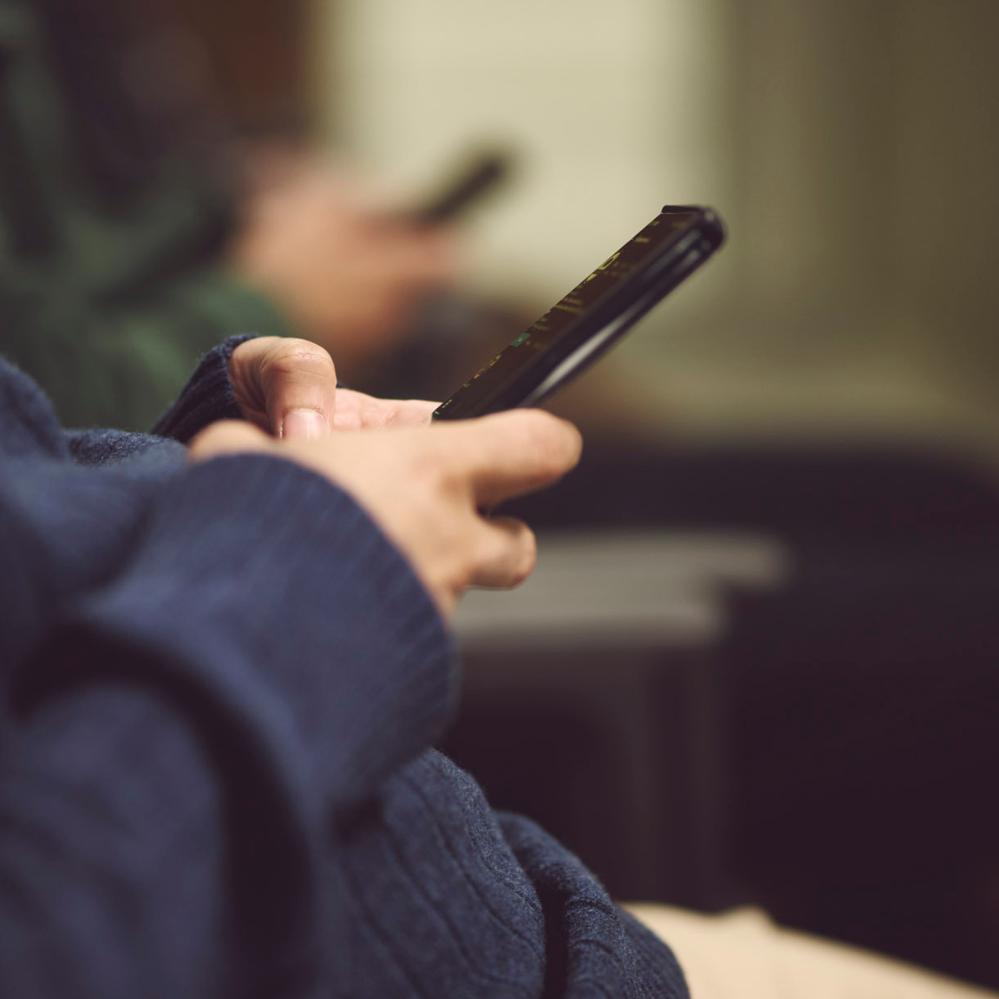 Two hands holding a mobile phone, with another pair of hands holding a phone out of focus in the background of a waiting room.