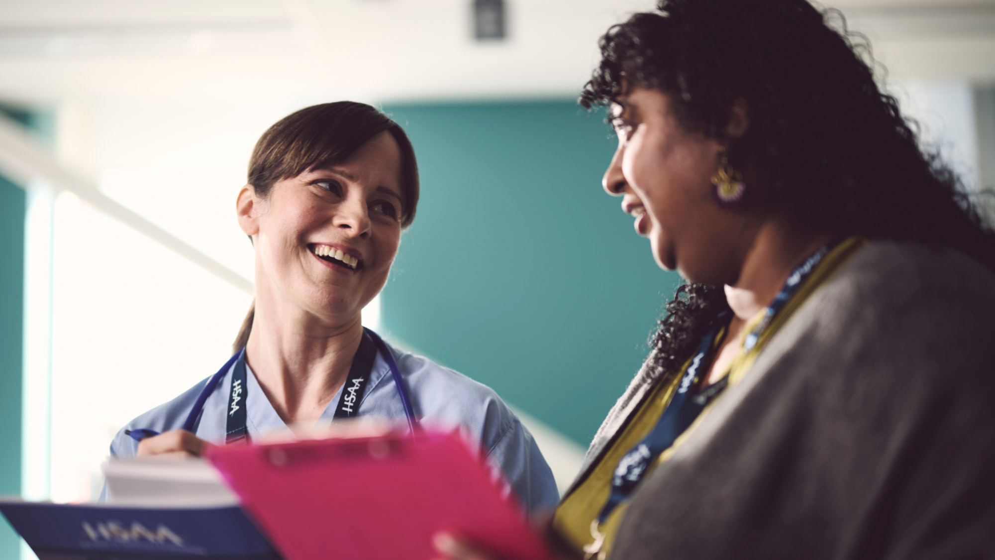 A white woman in scrubs and a brown woman in casual clothes talking to each other and smiling. Both are holding union paperwork.