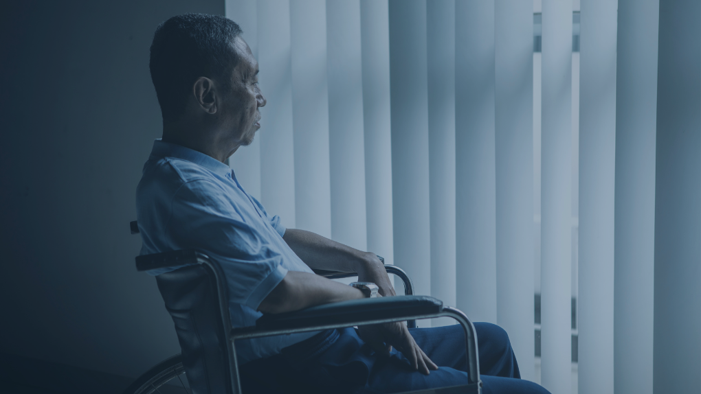 A patient sits in a wheelchair staring out a window through slatted blinds.