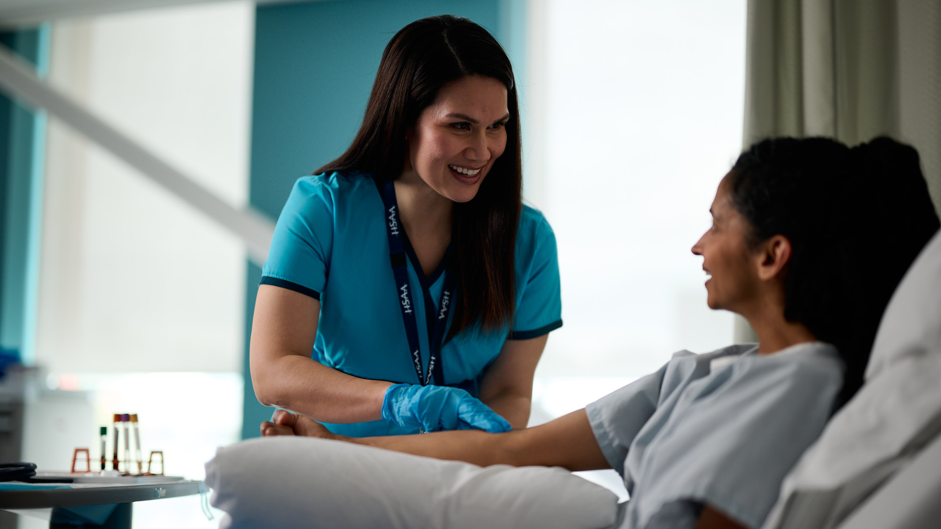 A patient sitting up in a hospital bed with an arm extended, and a lab tech by their side checking for a vein.