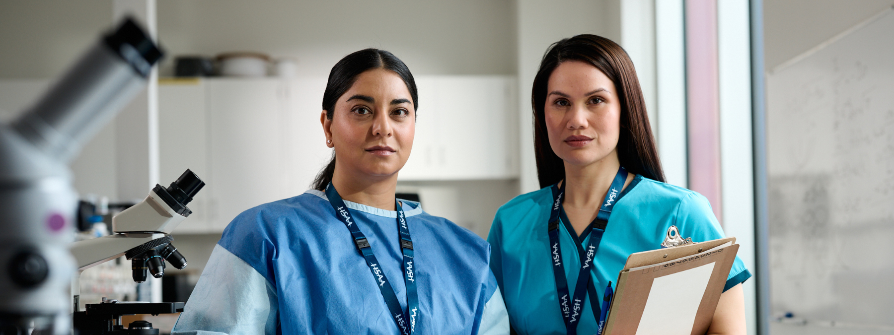 Two women, identified as HSAA members from their lanyards, stand in a medical lab with stern faces.