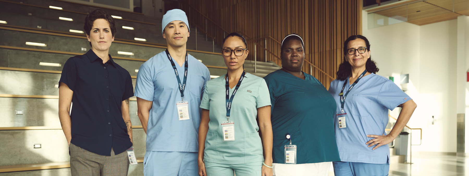 Five health care professionals in work appropriate attire stand in an open space, looking seriously at the camera. 