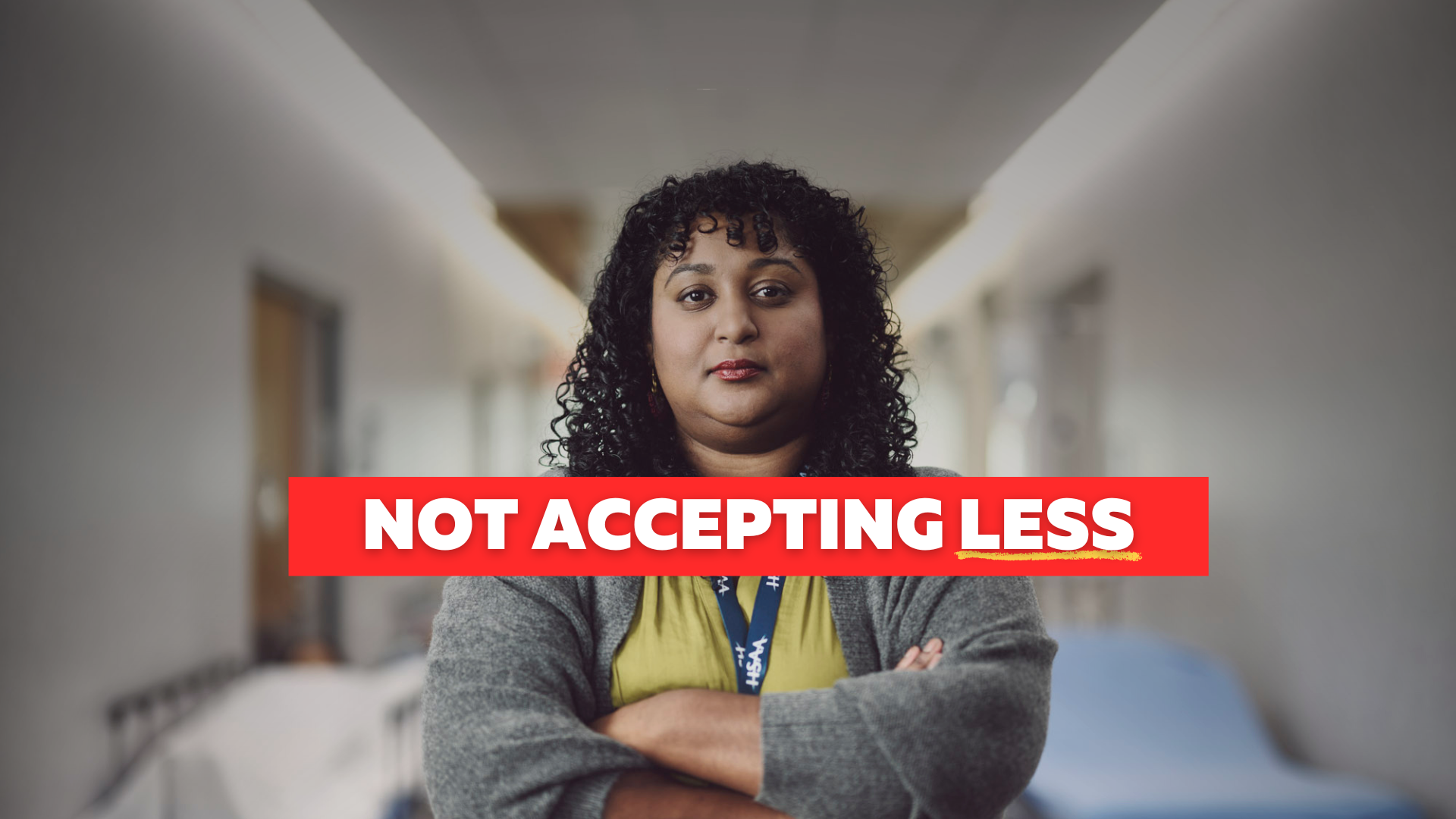A woman with brown skin and dark curly hair stands in a hospital hallway with her arms cross and a serious expression.