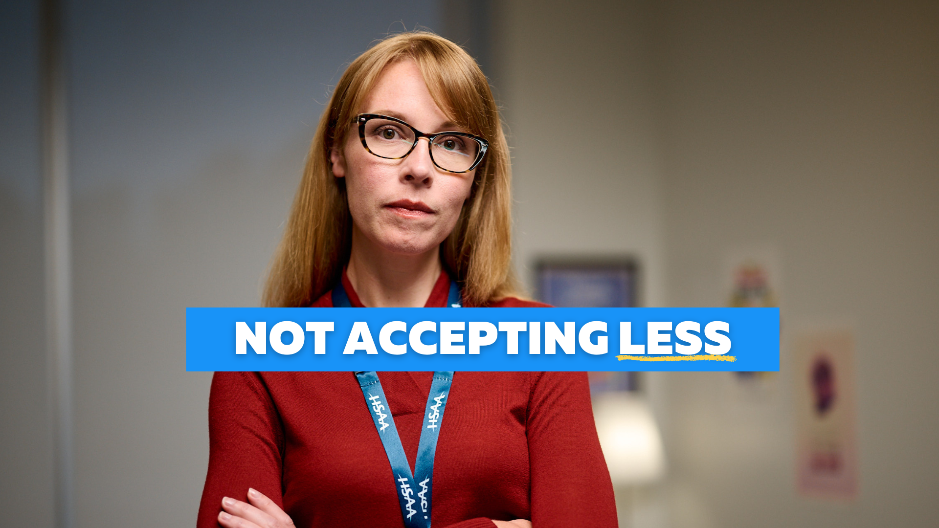 A woman with strawberry blonde hair wearing glasses and a red shirt stand with her arms crossed and a serious expression.