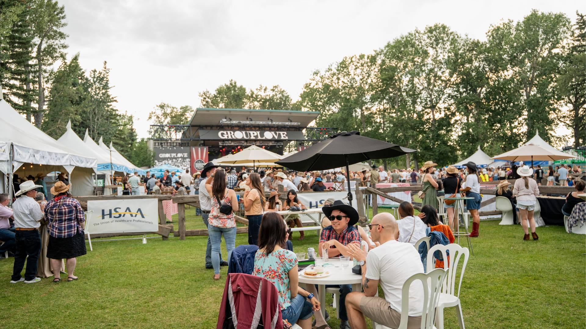 Wide shot of an outdoor event with tents and a concert stage and people sitting at tables or standing around at the event.