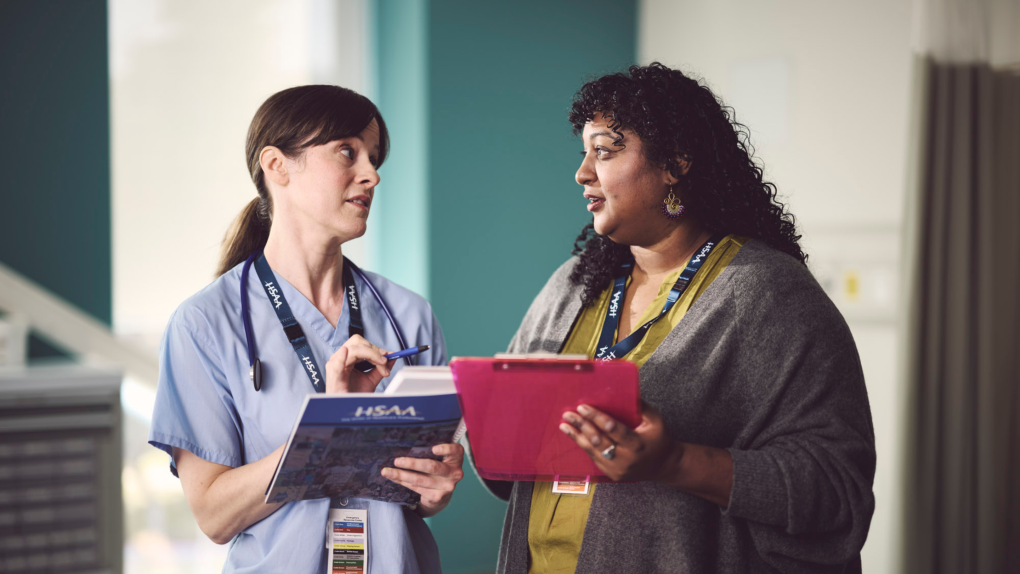 A white woman in scrubs and a brown woman in casual clothes talking to each other. Both are holding paperwork.